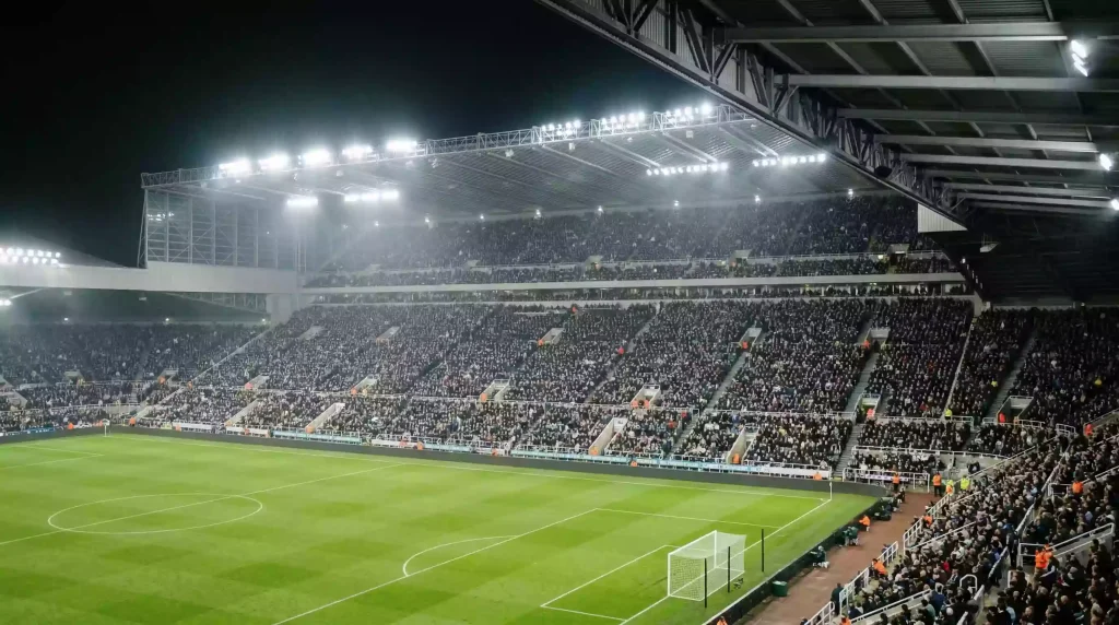Asistencia a la Premier League: tribuna de estadio inglés repleta de aficionados bajo focos nocturnos